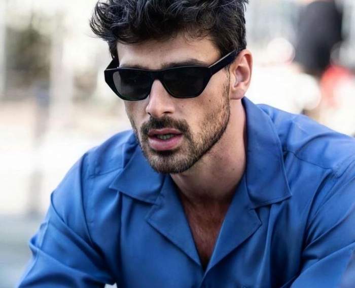Michele Morrone poartă ochelari de soare negri. Actorul e îmbrăcat cu o cămașă albastră.