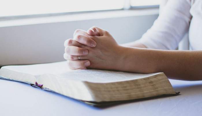 O carte sfântă este deschisă pe masă. O persoană ține mâinile pe ea și are degetele împreunate pentru rugăciune.