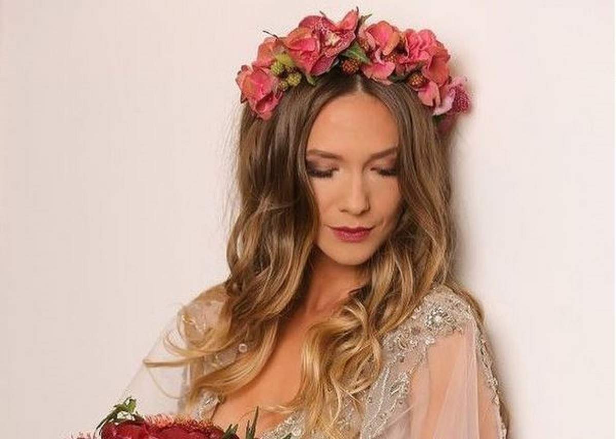Adela Popescu poartă o rochie albă cu brațe din voal și are pe cap o coroniță de flori roz. Vedeta privește în jos.