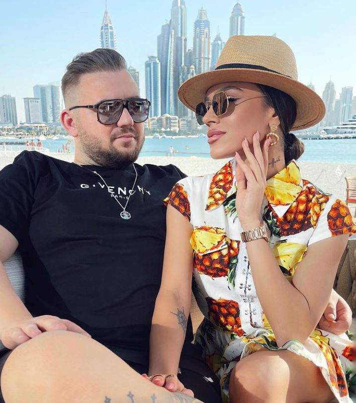 Karmen Simionescu este la o terasa in Dubai cu Bogdan Caplescu, ea poarta o rochie scurta cu palarie si ochelari de soare, el poarta haine negre si ochelari de soare