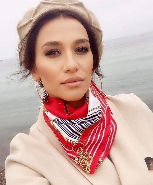 Claudia Pătrășcanu e la mare. Vedeta poartă o beretă crem pe cap. Artista e îmbrăcată cu un palton crem și are la gât o eșarfă roșie cu alb, nengru și auriu.