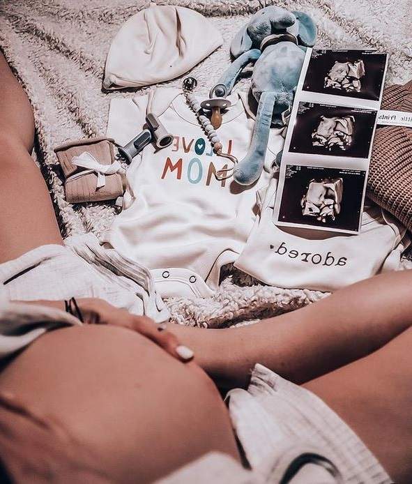 Flavia Mihășan își ține mâinile pe burtica de gravidă. Vedeta stă pe o pătură crem, iar lângă ea sunt haine de bebeluș și ecografia lui.
