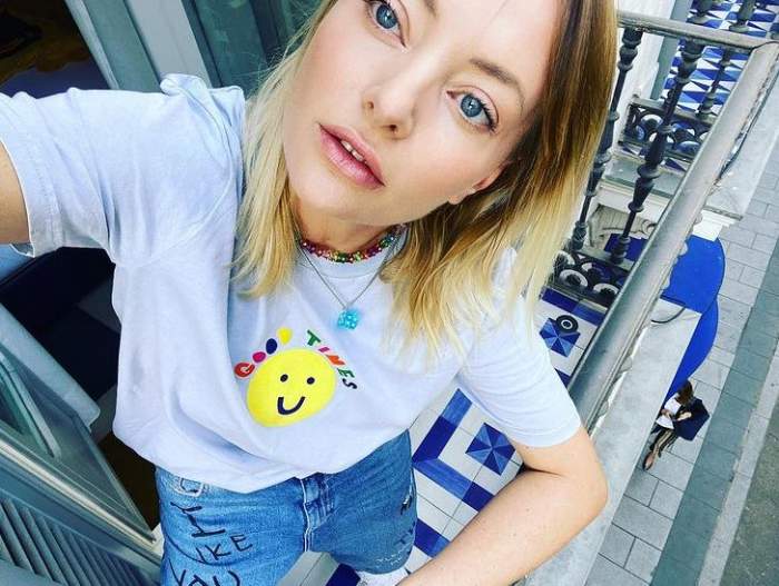 Delia Matache își face un selfie. Artista poartă un tricou alb cu un emoticon galben la nivelul pieptului și blugi albaștri.