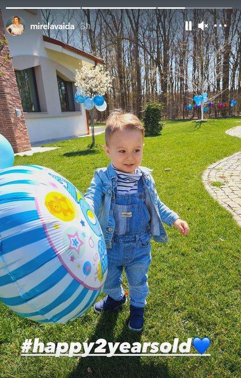 Tudor, fiul cel mic al Mirelei Vaida, se joacă cu un balon bleu în curtea casei. Copilul zâmbește și stă pe iarbă, purtând o salopetă din denim bleu.