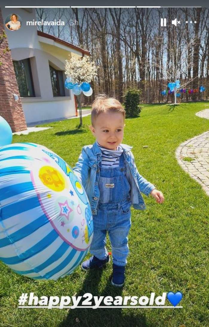 Tudor, fiul cel mic al Mirelei Vaida, se joacă cu un balon bleu în curtea casei. Copilul zâmbește și stă pe iarbă, purtând o salopetă din denim bleu.