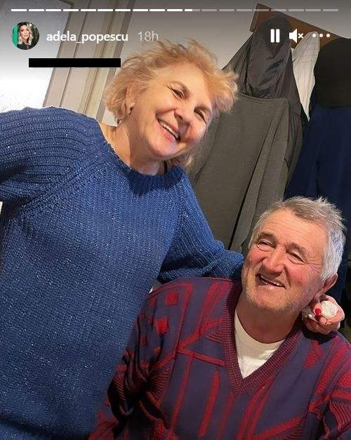 Părinții Adelei Popescu. Mama ei poartă un pulover albastru și își ține o mână pe umărul soțului ei, iar el poartă un pulover colorat în roșu și albastru. Amândoi zâmbesc.