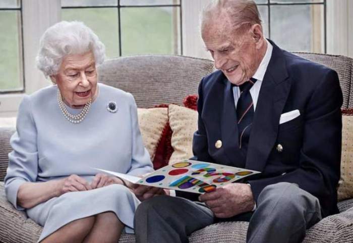 Regina Elisabeta a II-A și Prințul Philip stau pe o canapea gri. El ține în mână o carte din care citesc amândoi.
