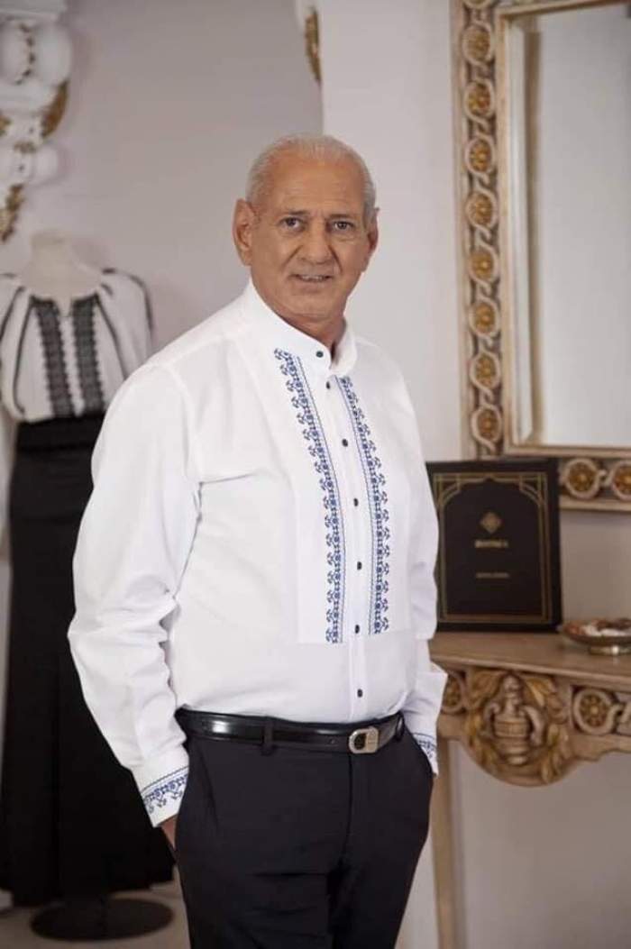 Gheorghe Zamfir poartă o cămașă albă, stil ie, cu model albastru și pantaloni negri. Artistul își ține mâinile în buzunare.