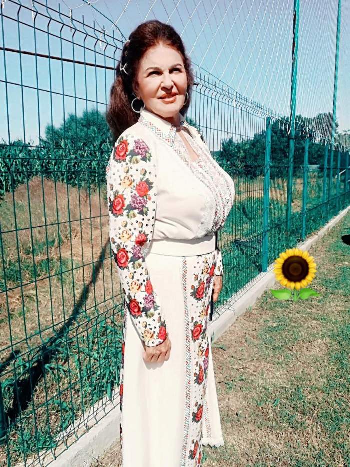Irina Loghin poartă rochie albă cu motive tradiționale colorate. Artista se află lângă un gard verde.