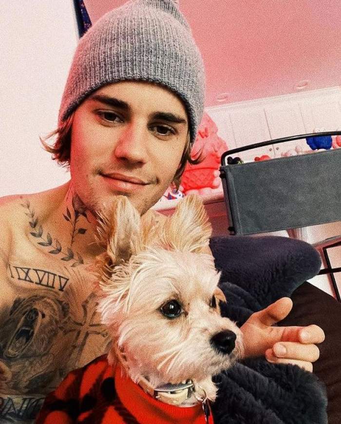 Justin Bieber poartă o căciulă gri. Artistul își ține în brațe câinele alb și zâmbește.