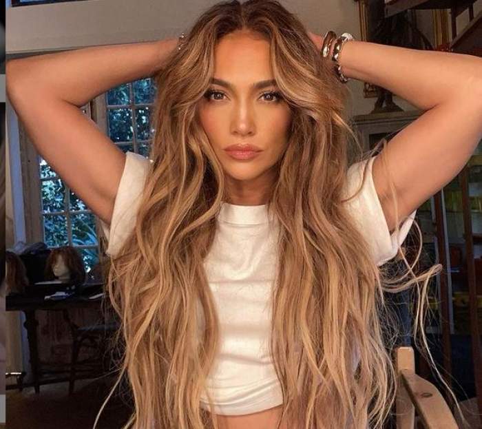 Jennifer Lopez poartă un tricou alb. Vedeta își ține mâinile ridicate la nivelul capului, după cap.