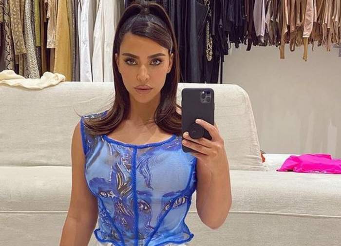 Kim Kardashian poartă un maiou albastru. Vedeta își face o poză în oglindă, cu telefonul, și are părul prins în coadă. În spatele ei se văd mai multe haine.