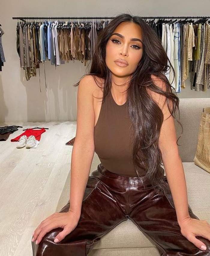 Kim Kardashian poartă un maiou maro și pantaloni din piele într-o nuanță mai închisă. Vedeta stă pe o canapea gri, iar în spatele ei sunt mai multe haine.