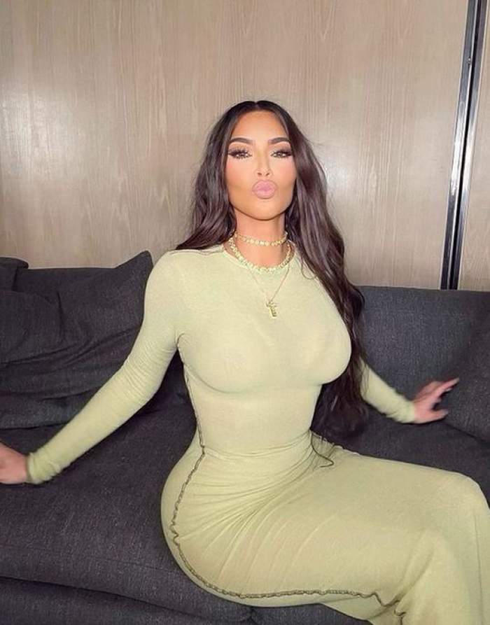 Kim Kardashian poartă o rochie mulată, verde. Vedeta stă pe o canapea gri și ține buzele țuguiate.