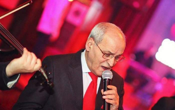 Nelu Ploieșteanu se află la un eveniment. Artistul cântă la microfon, poartă costum negru, iar pe dedesubt are o cămașă albă și cravată roșie.