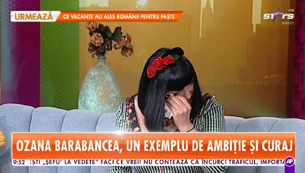 Ozana Barabancea poartă o rochie în dungi albe și negre. Artista are pe cap o coroniță roșie cu flori și își șterge lacrimile cu un șervețel.