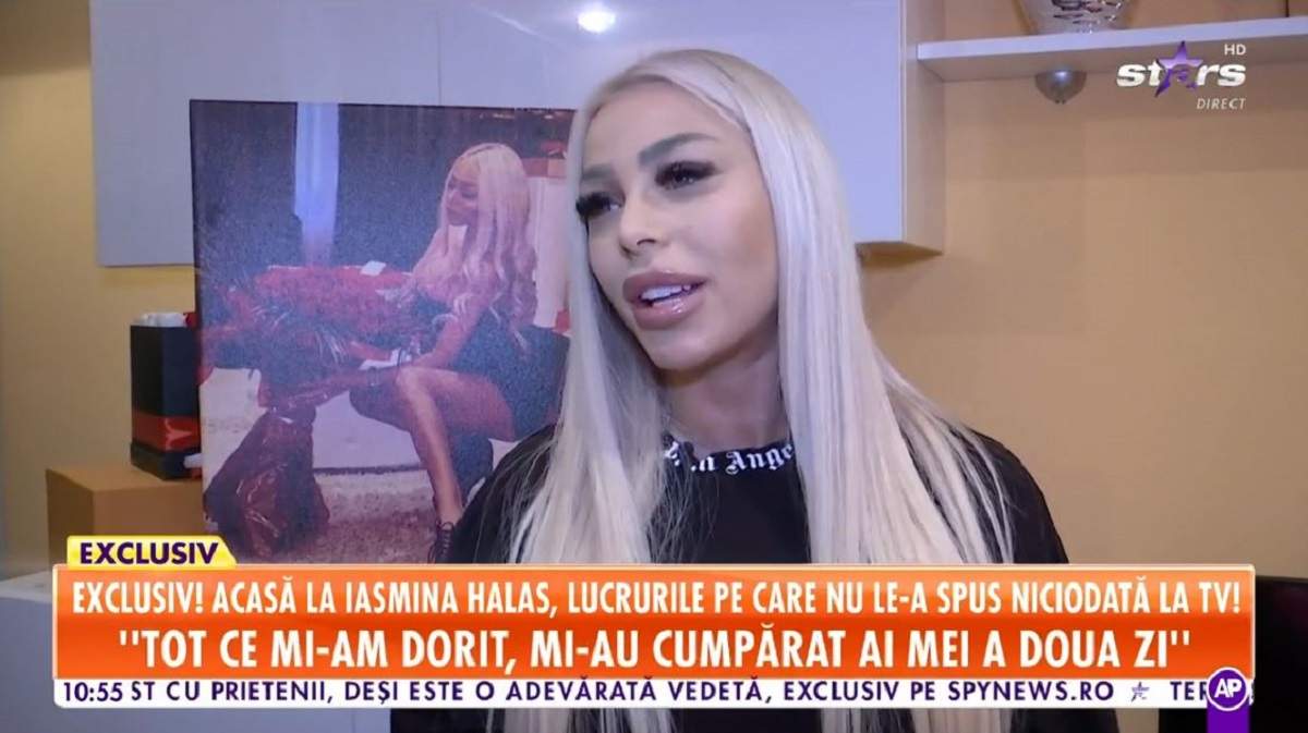 Iasmina Halas dă un interviu la Antena Stars. Vedeta poartă un tricou negru. În spatele ei  e un tablou cu ea în care ține un buchet de trandafiri în mână.