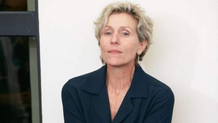 Frances McDormand poartă un sacou negru. Actrița e lipită cu spatele de un perete.