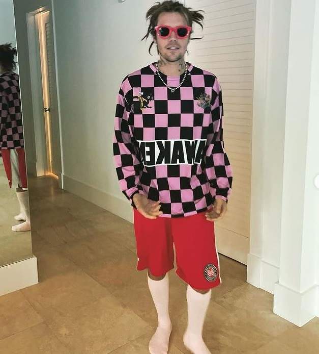 Justin Bieber poartă ochelari de soare cu rame roșii. Cântărețul poartă șosete albe, pantaloni scurți roșii și bluză în pătrățele negre și roz.