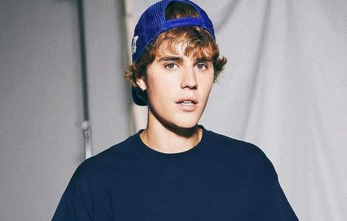 Justin Bieber poartă un tricou bleumarin și are o șapcă întoarsă pe cap, albastră. Artistul are gura întredeschisă.