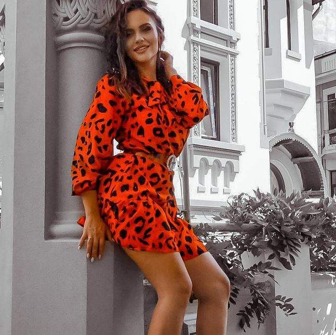 Cristina Șișcanu poartă o rochie oranj spre roșu, tip animal print. Vedeta zâmbește și se sprijină de un zid.