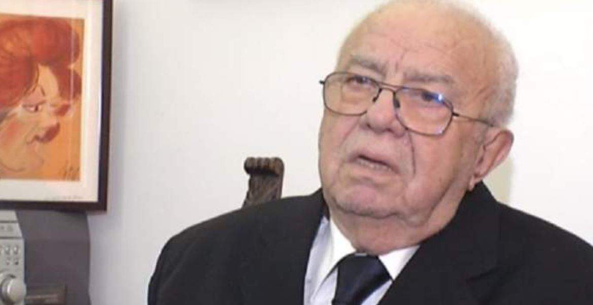 Alexandru Arșinel stă pe scaun. Artistul poartă ochelari de vedere și sacou negru.