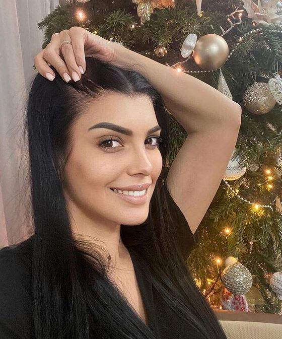 Andreea Tonciu se află în fața bradului de Crăciun. Vedeta poartă un tricou negru și zâmbește larg, ținându-și părul cu o mână.