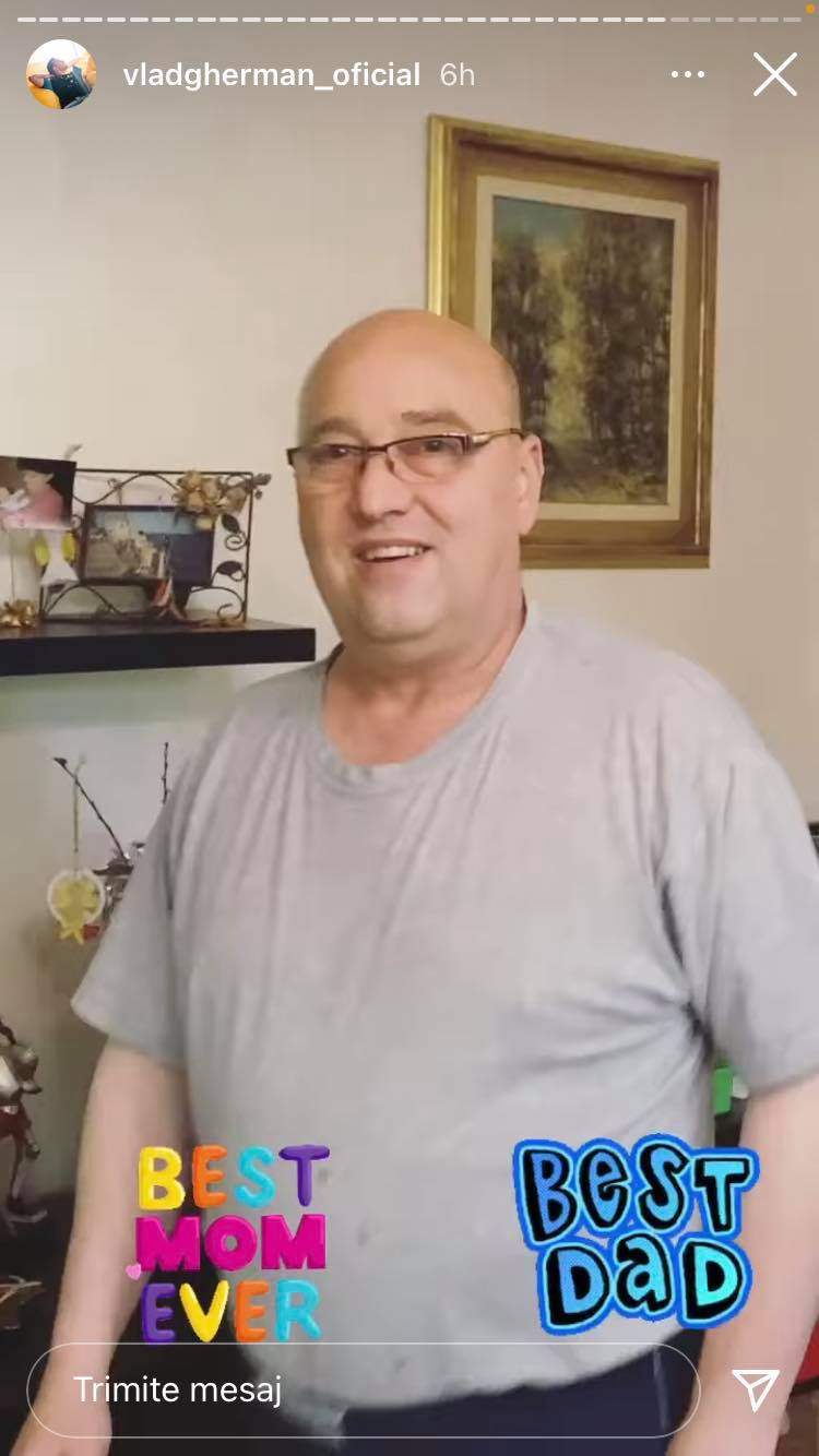 Tatăl lui Vlad Gherman poartă ochelari de vedere și un tricou gri. Bărbatul zâmbește, iar în spatele lui sunt mai multe tablouri.