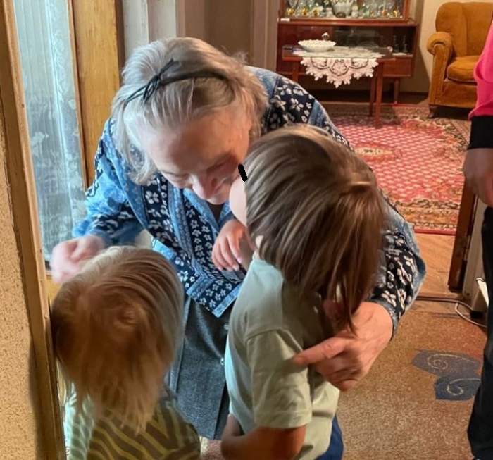 Băieții Adelei Popescu și cu străbunica lor se află în casă. Femeia poartă o cămașă colorată în negru și bleu, cu floricele, și fustă gri și îi cuprinde pe copii în brațe. Micuții poartă tricouri verzi.
