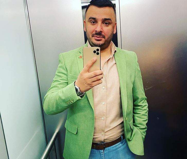 Liviu Guță e în lift. Artistul poartă cămașă crem, sacou verde și blugi albaștri.
