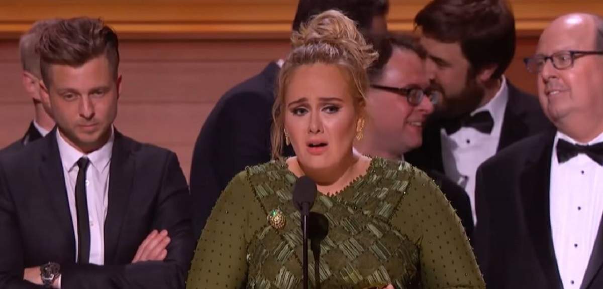 Adele la premiile Grammy. Vedeta ține un discurs în lacrimi și poartă o rochie verde. În spatele ei sunt mai mulți bărbați.