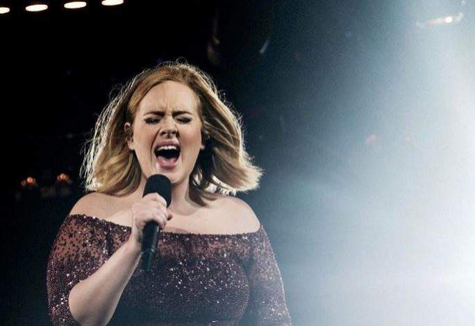 Adele cântă la microfon, pe scenă. Artista poartă o rochie sclipitoare.