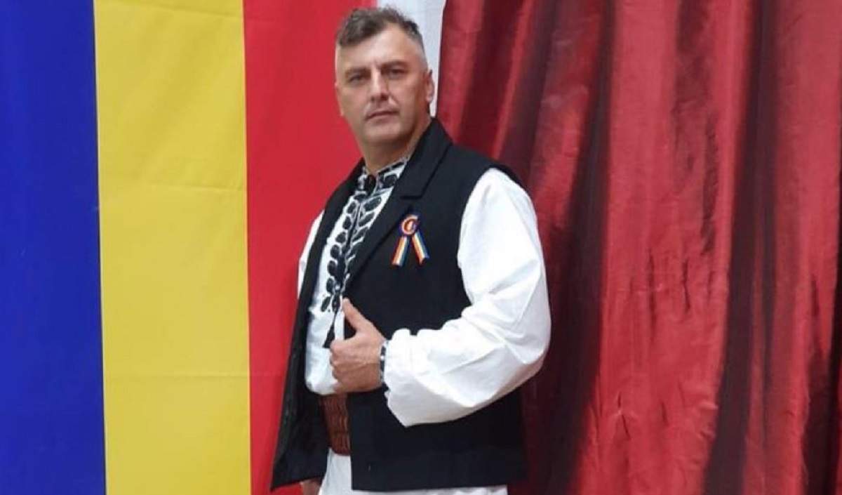 Corin Dobrinescu poartă costum popular, cu ie albă și veste neagră. În spatele lui se află steagul României.
