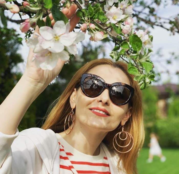 Alina Sorescu poartă ochelari de soare. Vedeta ține mâna în florile unui copac.