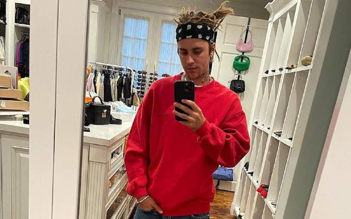 Justin Bieber poartă o bluză roșie și blugi albaștri și își face o poză cu telefonul în oglindă.