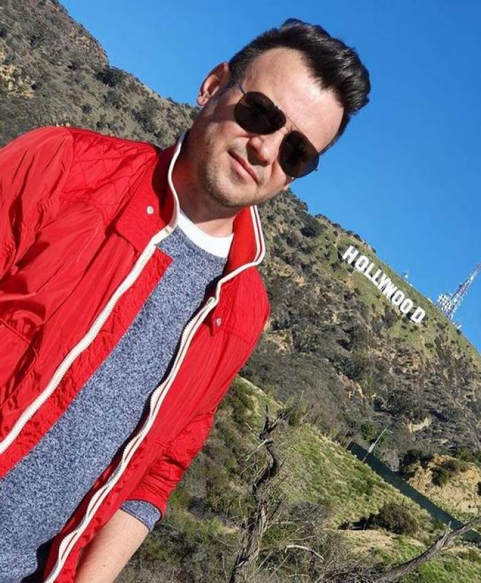 Dan Deneș poartă ochelari de soare, bluză gri, iar pe deasupra o altă bluză roșie, descheiată.