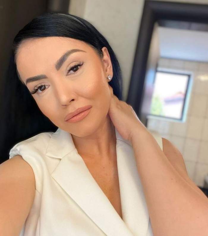 Andreea Mantea își face un selfie și poartă un sacou alb, ținându-și o mână pe gât.