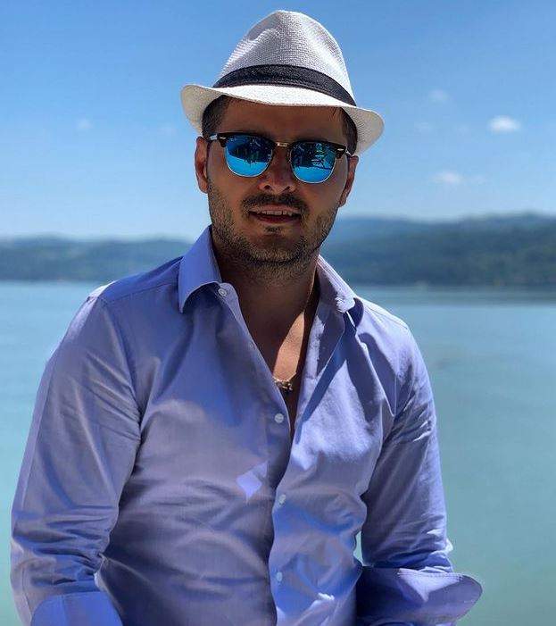 Liviu Vârciu se află la mare. Artistul poartă o pereche de ochelari de soare, o pălărie albă cu dungă neagră și o cămașă bleu.
