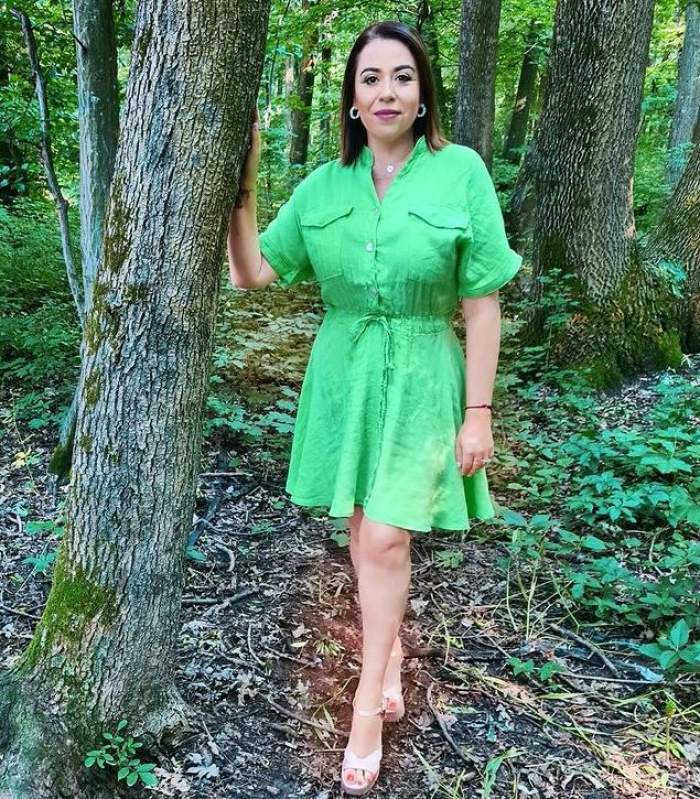 Oana Roman se ține cu o mână de un copac, în parc. Vedeta poartă rochie verde și sandale albe.