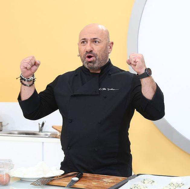 Cătălin Scărlătescu poartă o uniformă de bucătar neagră. Chef-ul e în bucătăria de la Chefi la cuțite și ține pumnii ridicați în aer, având gura deschisă.