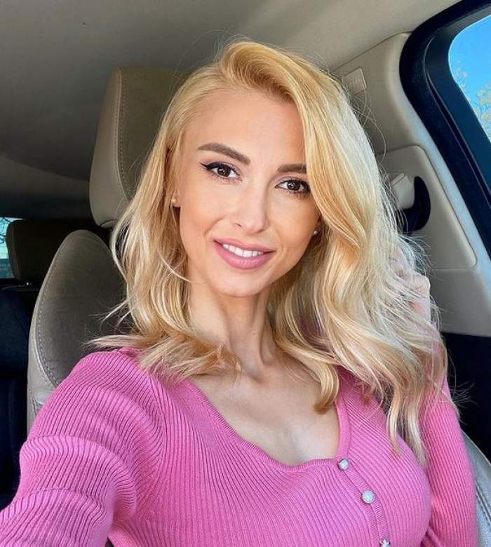 Andreea Bălan e în mașină și poartă o bluză roz. Artista zâmbește larg.