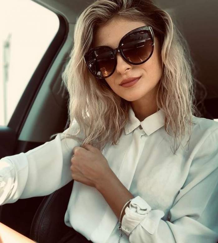 Elena Matei își face un selfie din mașină purtând ochelari de soare și cămașă albă. Tânăra zâmbește slab.
