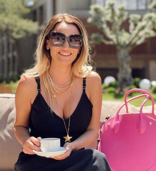 Carmen Neogiță e la terasă, ține o ceașcă albă de cafea în mână și poartă ochelari de soare și rochie neagră.