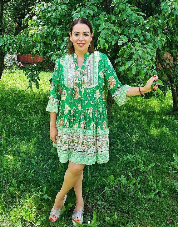 Oana Roman e în grădină, poartă rochie verde cu imprimeu colorat și zâmbește. În spatele ei sunt copaci.
