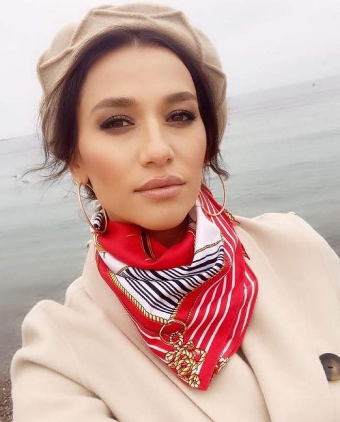 Claudia Pătrășcanu e la mare. Vedeta poartă o beretă crem pe cap. Artista e îmbrăcată cu un palton crem și are la gât o eșarfă roșie cu alb, nengru și auriu.