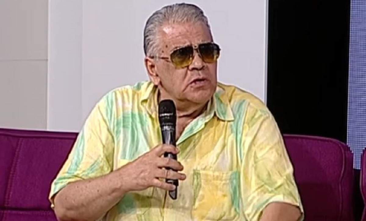 Florin Condurățeanu poartă ochelari de soare, vorbește la microfon și e îmbrăcat în cămașă cu mâneca scurtă galbenă cu model verde.