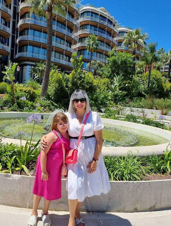 Oana Roman și fiica ei în vacanța din Franța. În spatele lor să văd mai multe clădiri, palmieri și o fântână artieziană. Cele două zâmbesc și se țin în brațe.