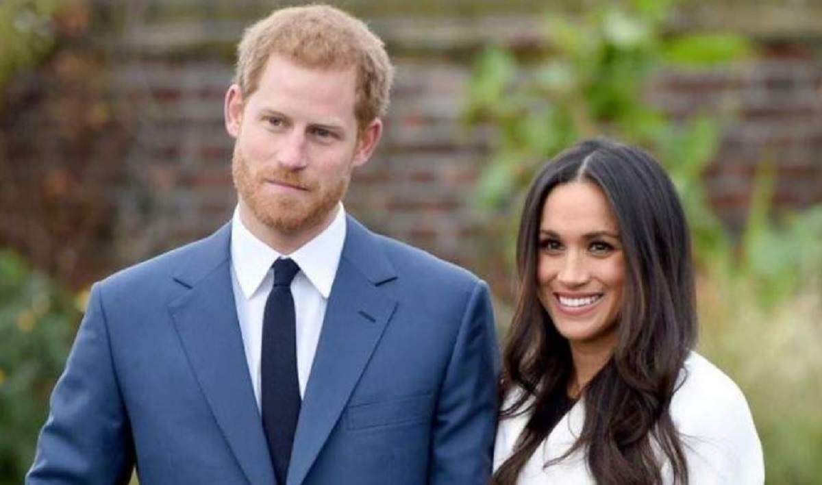 Meghan Markle și Prințul Harry se află afară. Ea poartă palton alb și zâmbește, iar el sacou albastru și cămașă albă.