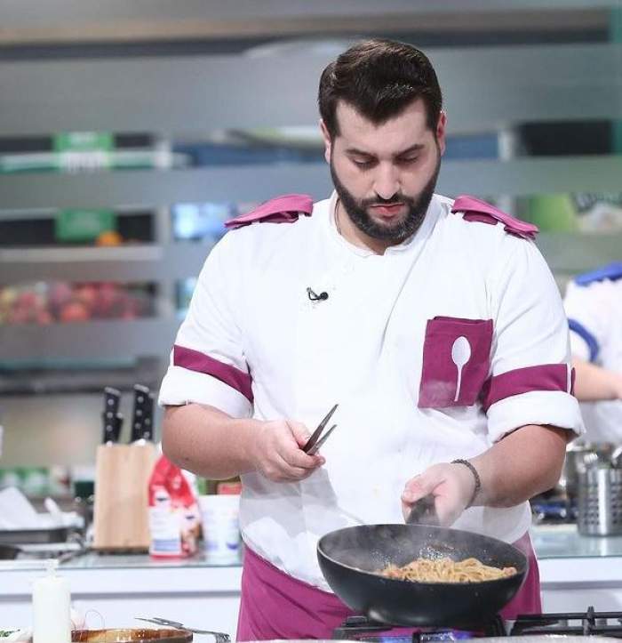 Enzo gătește la Chefi la cuțite, ținând în mână o ustensilă și tigaie neagră. Tânărul poartă uniformă albă cu model mov, de bucătar.