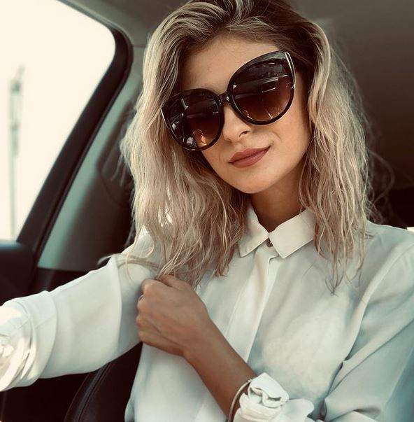 Elena Matei își face un selfie din mașină purtând ochelari de soare și cămașă albă.